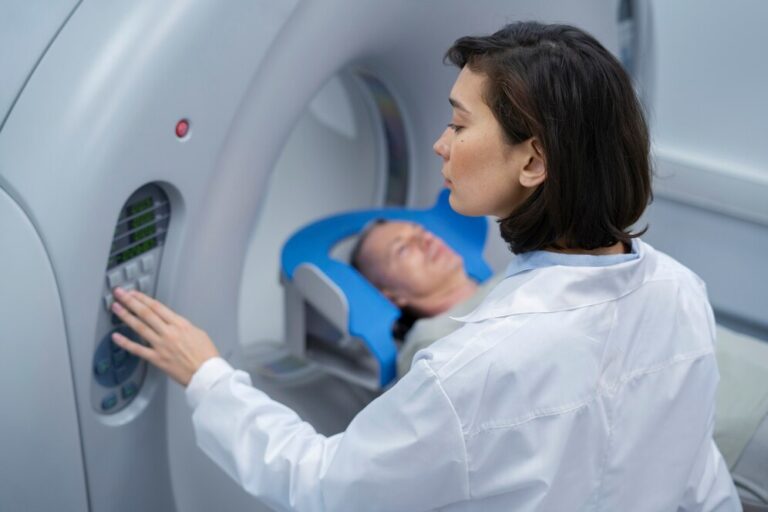 A foto mostra uma técnica de radiologia, uma mulher vestindo jaleco branco, operando um aparelho de ressonância magnética. Ela está ajustando os controles no painel do equipamento enquanto observa atentamente um paciente, que está deitado dentro do túnel do scanner, visível ao fundo.