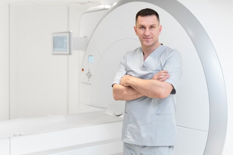 A imagem mostra um técnico de radiologia, um homem com uniforme cirúrgico cinza, de braços cruzados e com uma expressão séria e confiante. Ele está de pé em frente a um moderno aparelho de ressonância magnética.